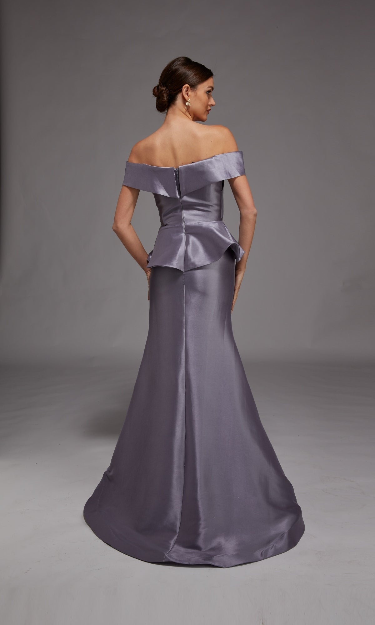 A woman stands facing away, wearing the Alyce 27827 long formal dress—a fitted, off-the-shoulder lavender mermaid gown with a peplum detail and smooth satin finish.